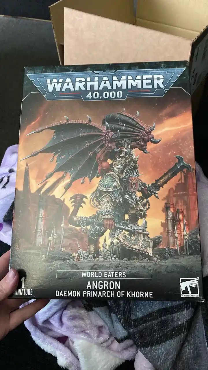 A hand holds a Warhammer 40,000 miniature box featuring Angron, Daemon Primarch of Khorne, a massive demonic warrior with…