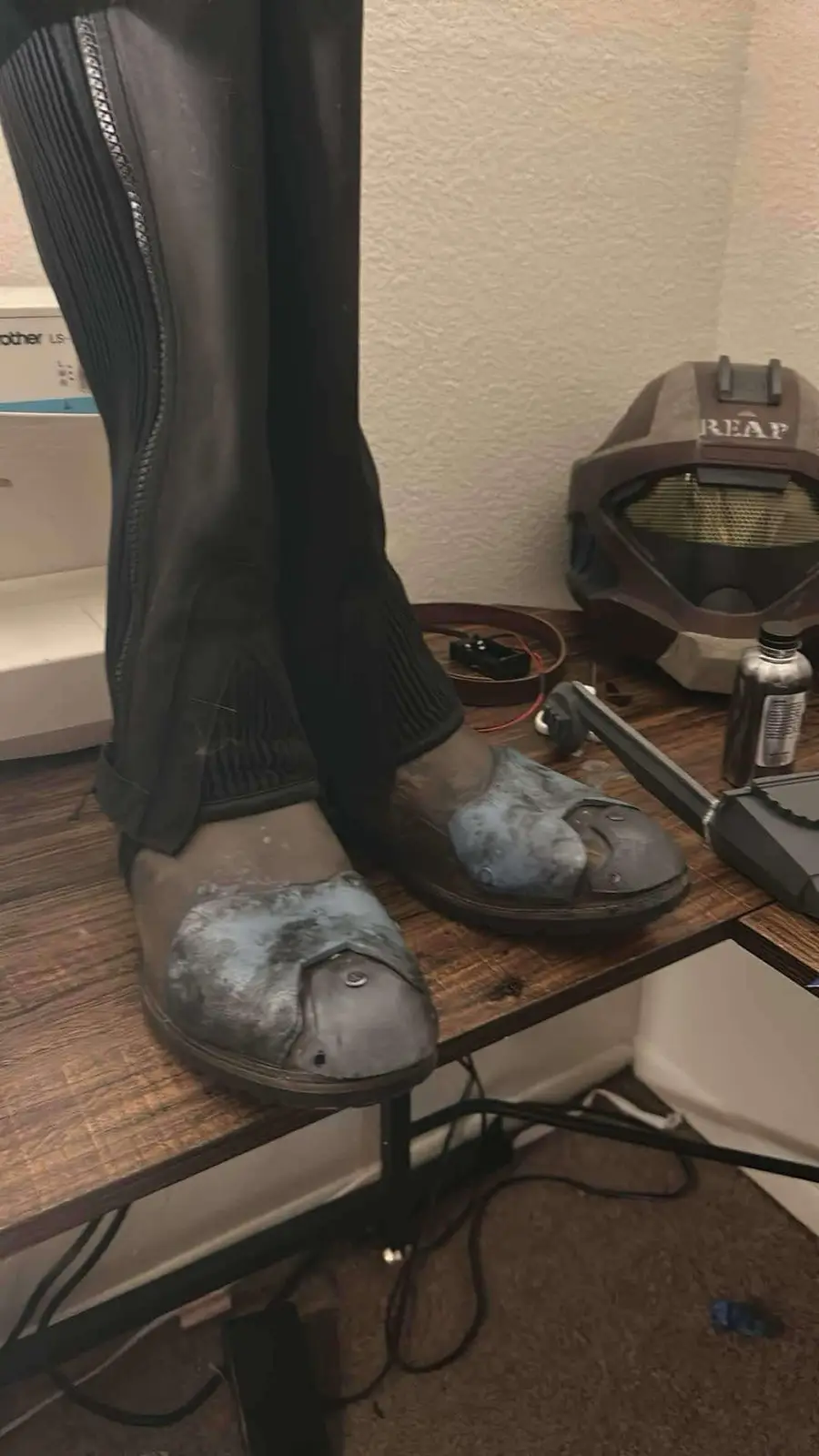 A close-up photo shows a person's legs in black riding boots standing on a wooden workbench. The boots have large…