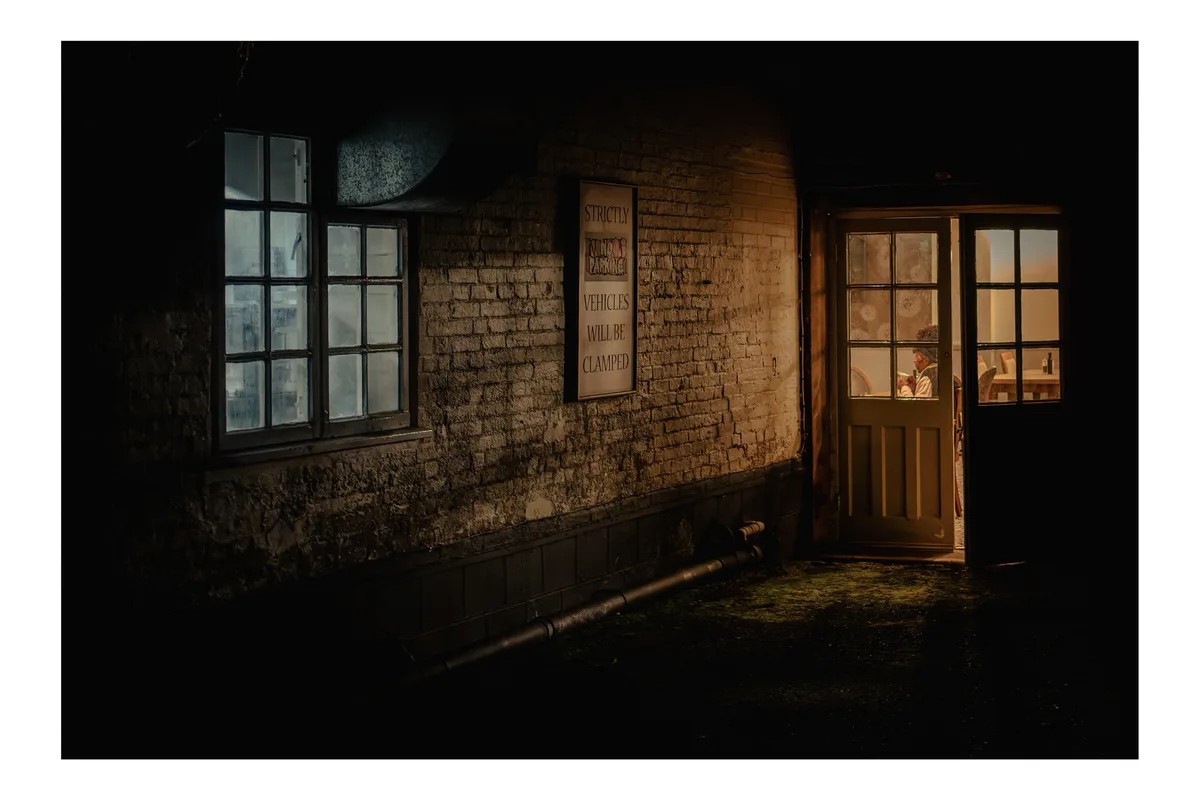 A dimly lit, atmospheric alleyway with a brick wall, a window, and a partially open door revealing a warmly lit interior. A…