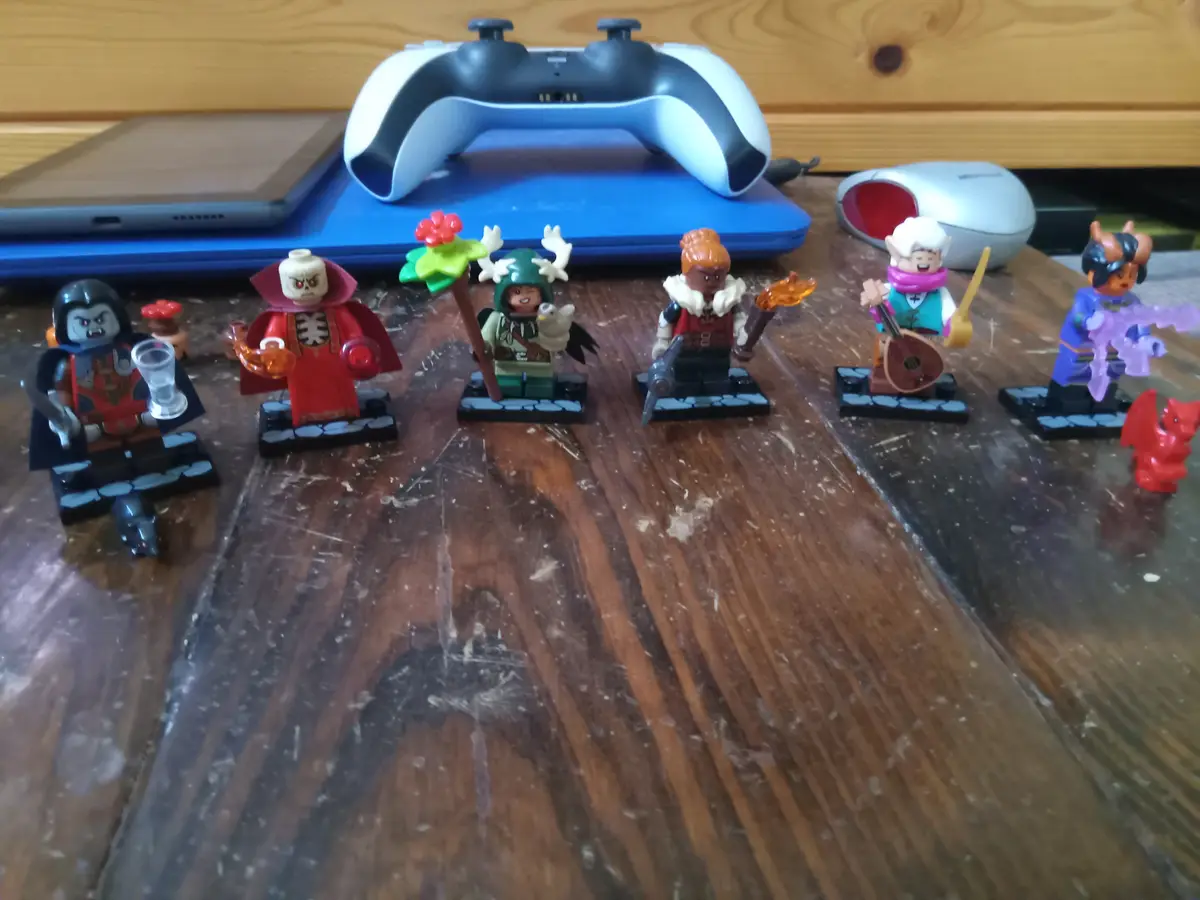 Six Lego minifigures representing fantasy characters are arranged in a row on a scratched wooden table, with a PlayStation 5…