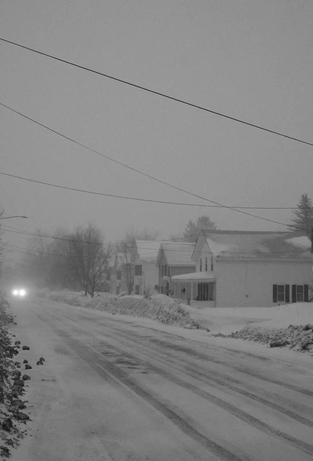 A black-and-white photo shows a snow-covered rural road with houses on the right and a car's headlights visible in the…