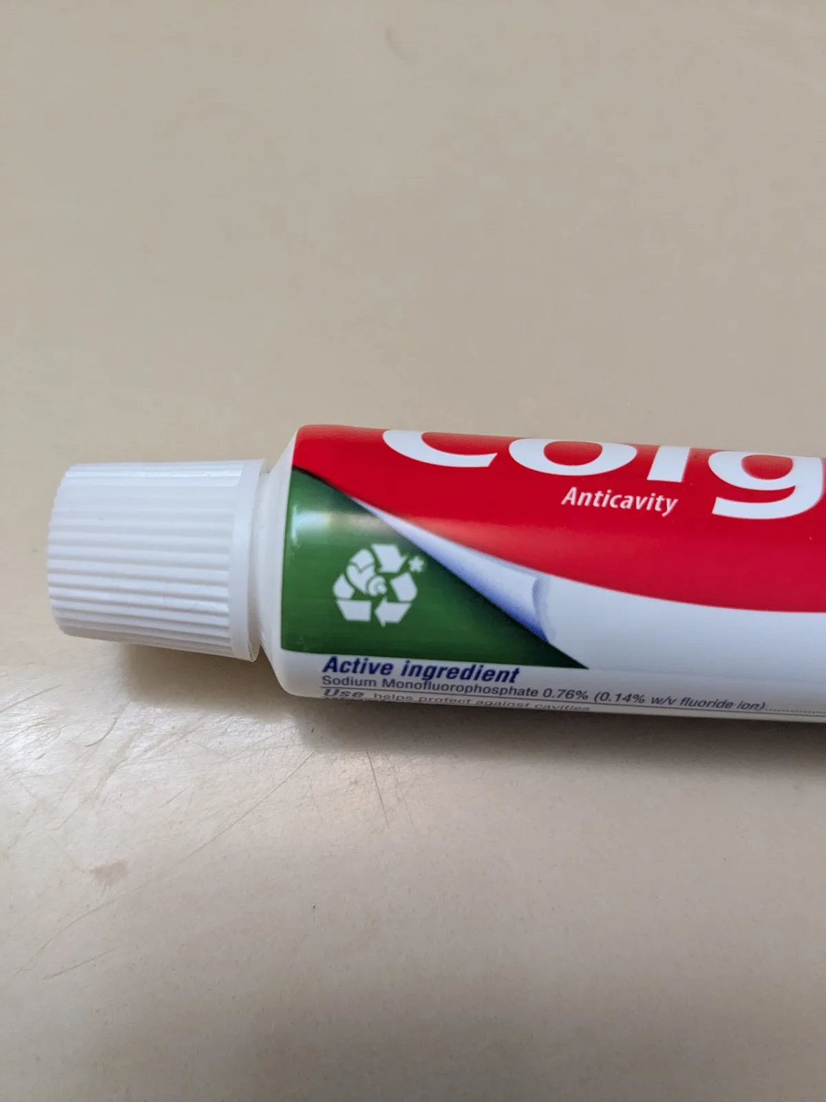 A close-up photo of a partially used Colgate Anticavity toothpaste tube lying on a scratched beige surface. The tube's label…