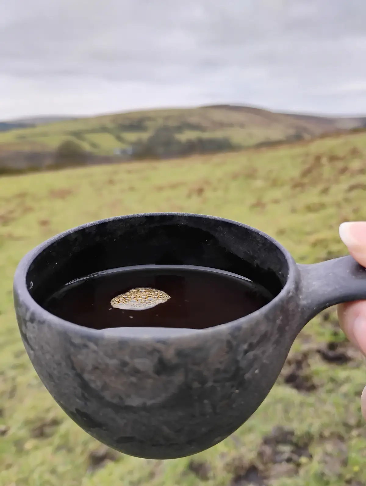 A hand holds a dark, textured ceramic mug filled with a dark liquid, likely coffee or tea, against a blurred background of…