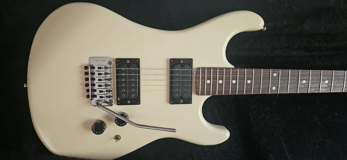 A cream-colored electric guitar with a Floyd Rose tremolo system, two humbucker pickups, and a dark fretboard with dot…