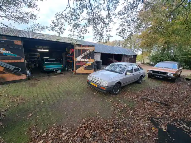 An outdoor shot of a cluttered, rustic garage with multiple vintage cars parked outside and inside. A silver sedan is in the…