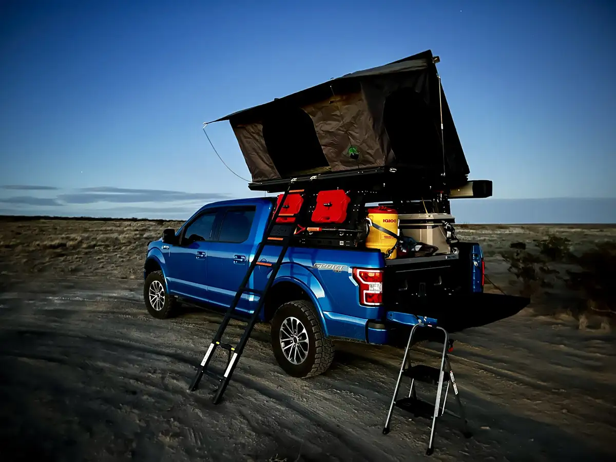 A blue pickup truck is parked in a desert landscape with a rooftop tent fully deployed and a ladder leaning against it. The…