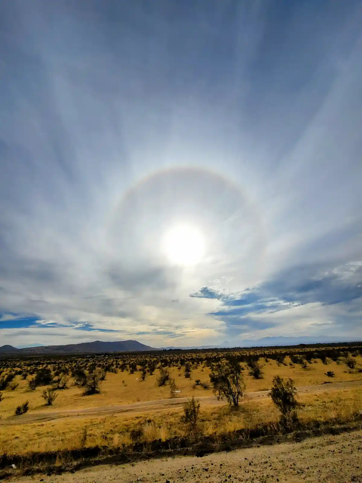 A wide-angle photo shows a bright sun in a partly cloudy sky, surrounded by a faint, circular halo of light. Below, a dry…