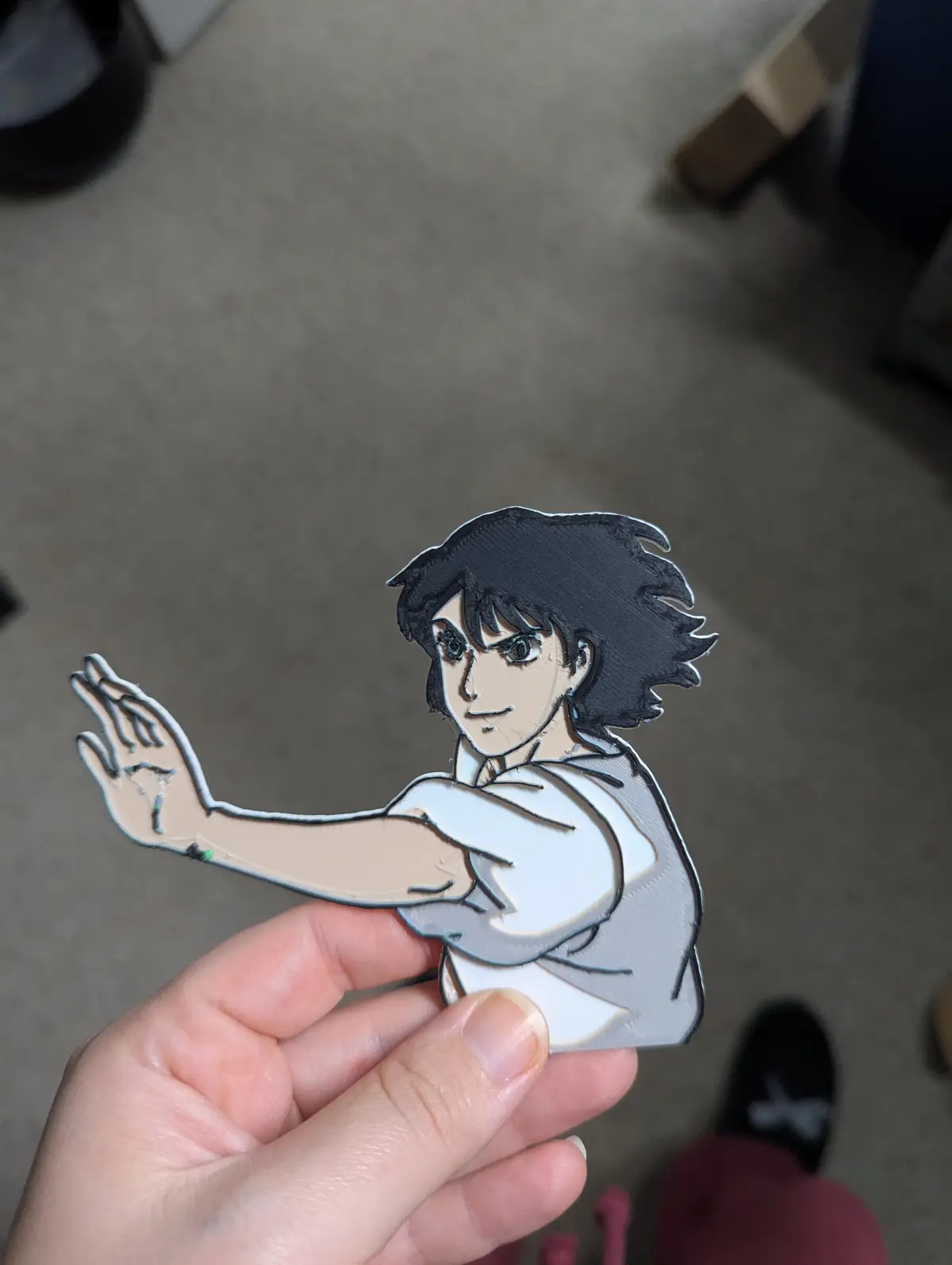 A hand holds a 3D-printed, painted cutout of the anime character L (from Death Note), showing his iconic pose with one hand…
