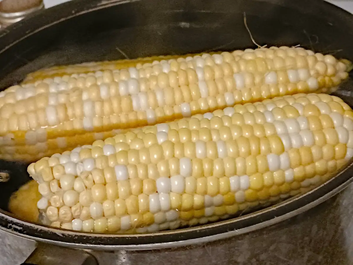 Two ears of yellow and white striped corn on the cob are shown steaming in a black frying pan. The overlaid text reads 'i'm…