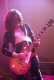 A musician with long curly hair, wearing a black jacket and pants, plays a sunburst electric guitar on stage under purple…