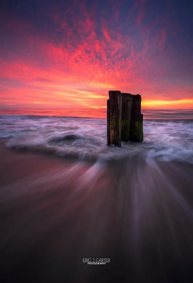 A long-exposure photograph shows a weathered wooden post standing in the ocean at sunset, with vibrant red and orange clouds…