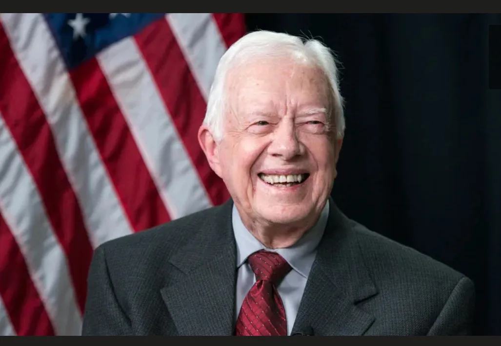 a smiling older man with white hair, wearing a dark suit and red tie, stands in front of an american flag. The overlaid text…