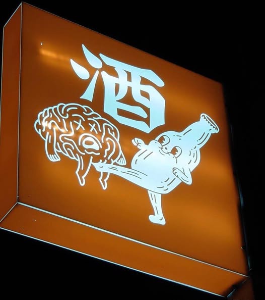 An illuminated orange sign features a large white Japanese kanji character '酒' (sake) above a cartoon bottle character…