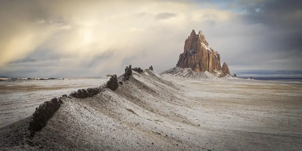 A wide, snowy desert landscape featuring a prominent, jagged rock formation under a cloudy sky with sunlight breaking…