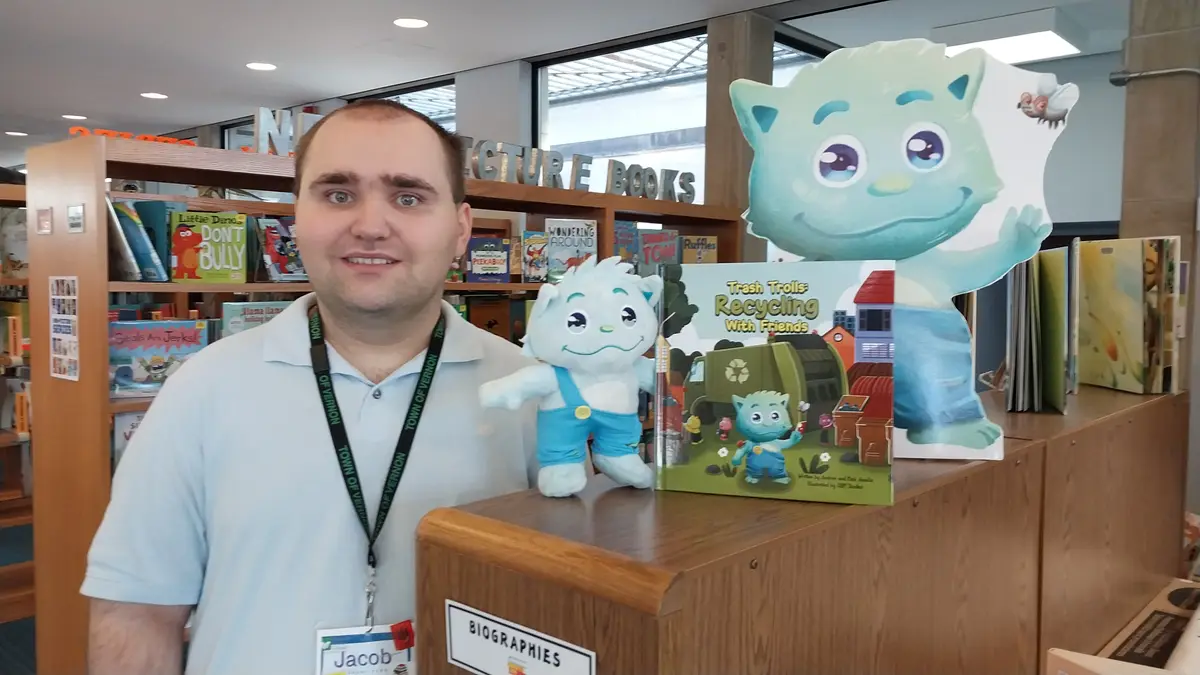 A man named Jacob, wearing a 'Town of Vernon' lanyard, stands in a library next to a display featuring a plush toy, a book…