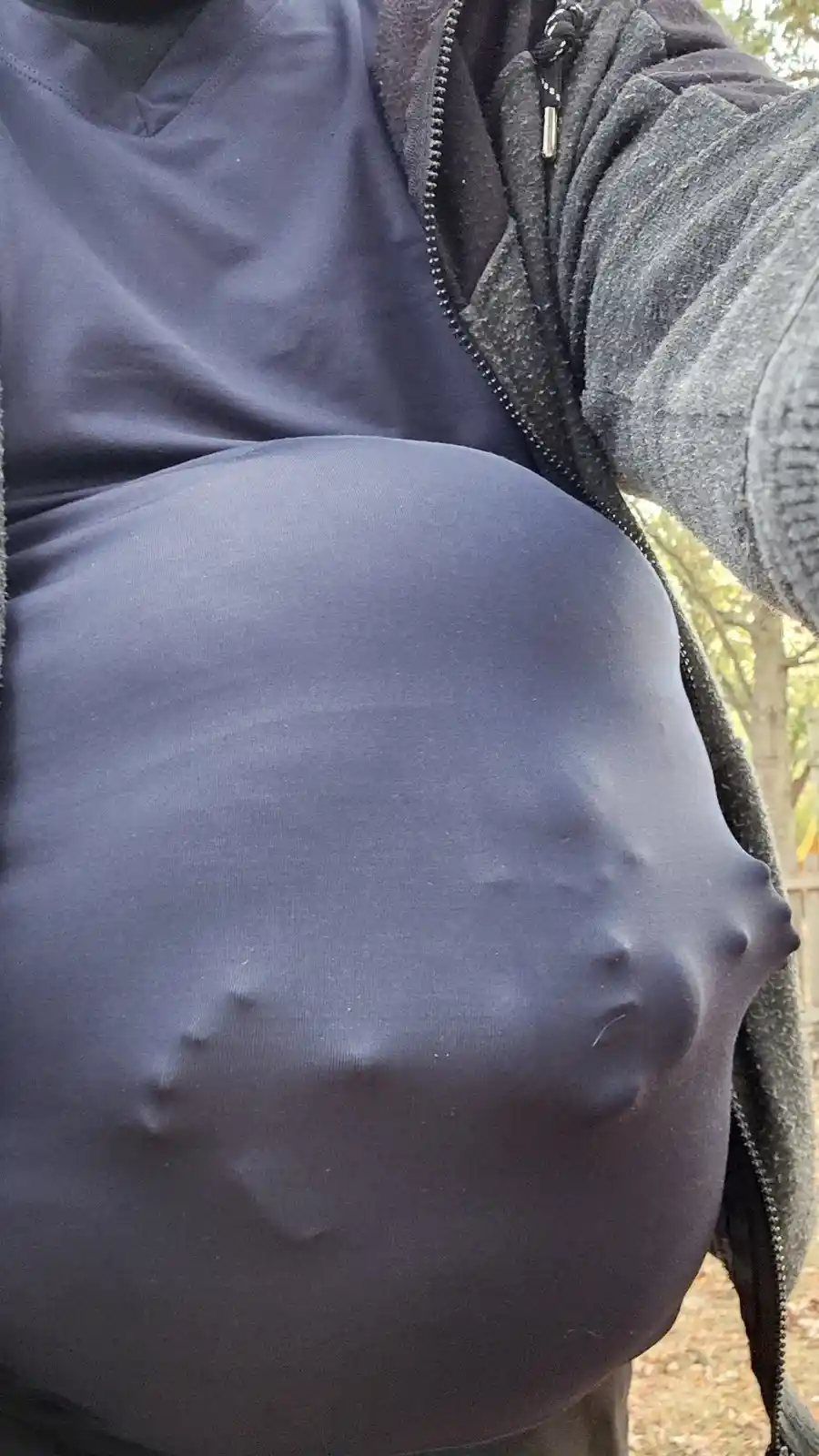 A close-up photo shows a person wearing a dark zip-up hoodie with the zipper partially open, revealing a very large, rounded…