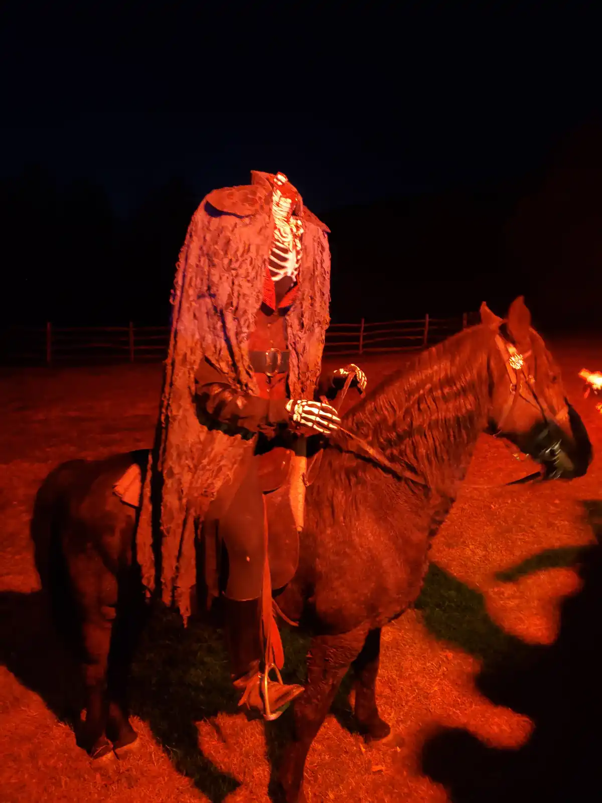 A person in a full skeleton costume, including a skull mask and ribcage torso, is riding a dark horse under red-orange…