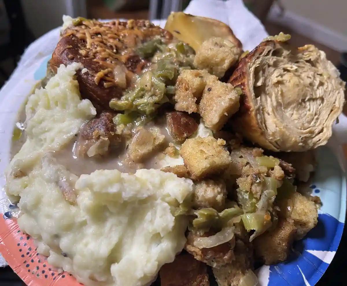A close-up photo shows a paper plate piled high with holiday food including mashed potatoes, stuffing with green beans and…