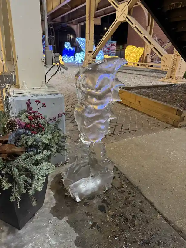 An outdoor photo shows a melting ice sculpture shaped vaguely like a person, standing on a wet sidewalk next to a potted…