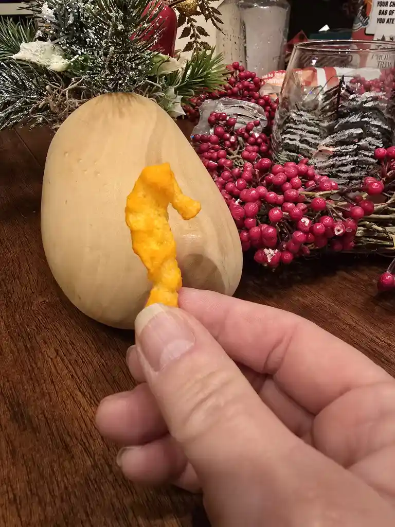 A hand holds a small, orange, seahorse-shaped cheese chip in front of a light-colored, egg-shaped squash. The background…
