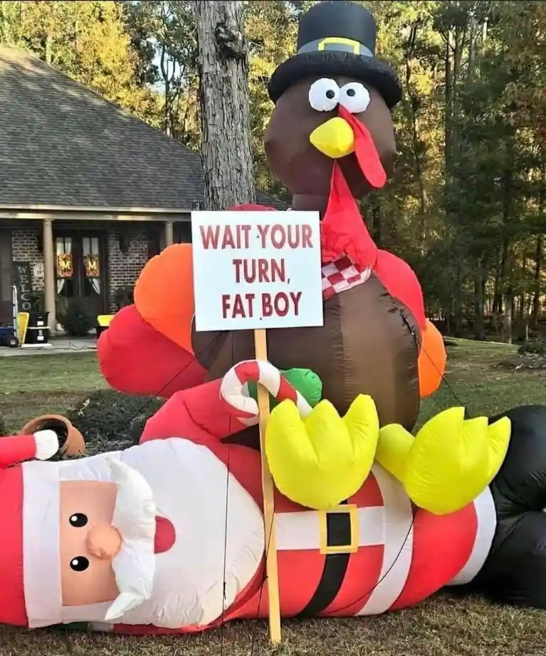 An inflatable Thanksgiving turkey wearing a pilgrim hat stands over a fallen inflatable Santa Claus holding a sign that…