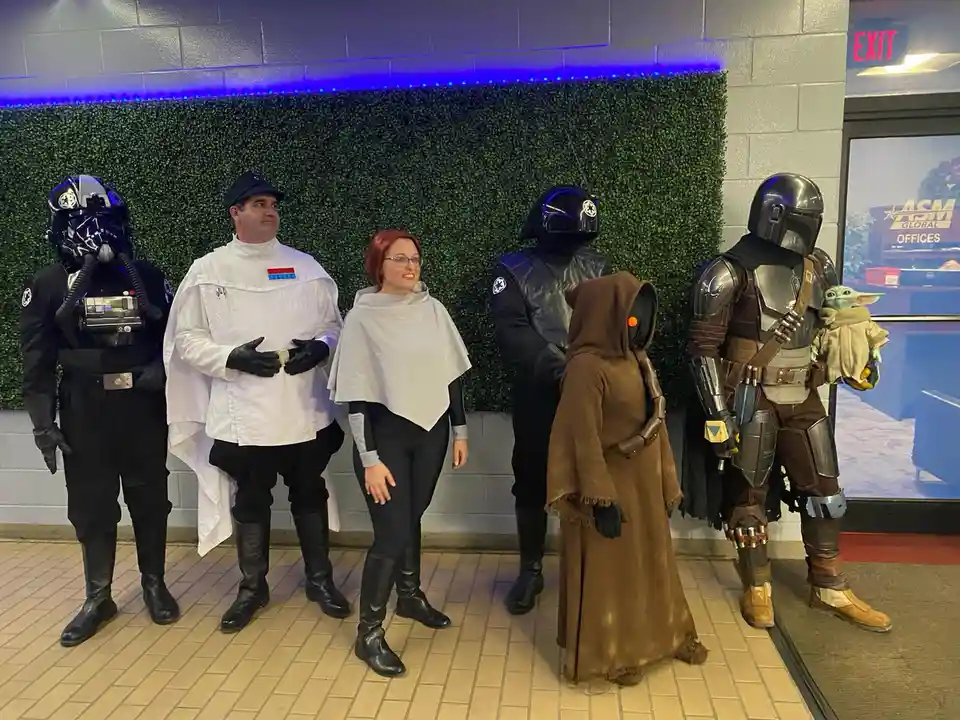 six people in Star Wars costumes stand in a line against a green hedge wall with blue LED lights above. From left to right…