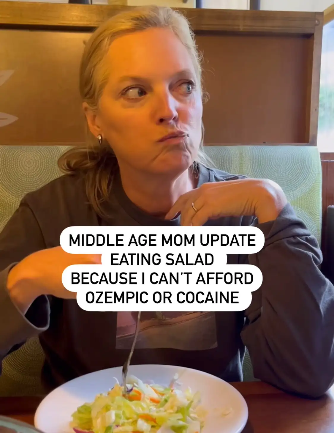 A middle-aged woman with blonde hair sits at a restaurant table, looking sideways with a skeptical, slightly disgusted…