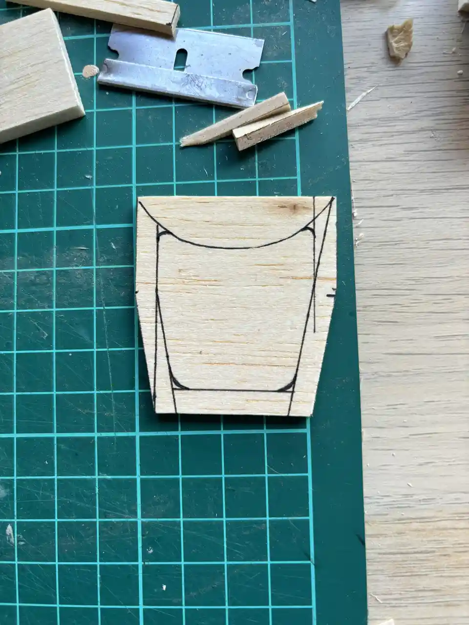 A piece of light-colored wood with a hand-drawn outline of a glass on it sits on a green cutting mat. Surrounding the wood…