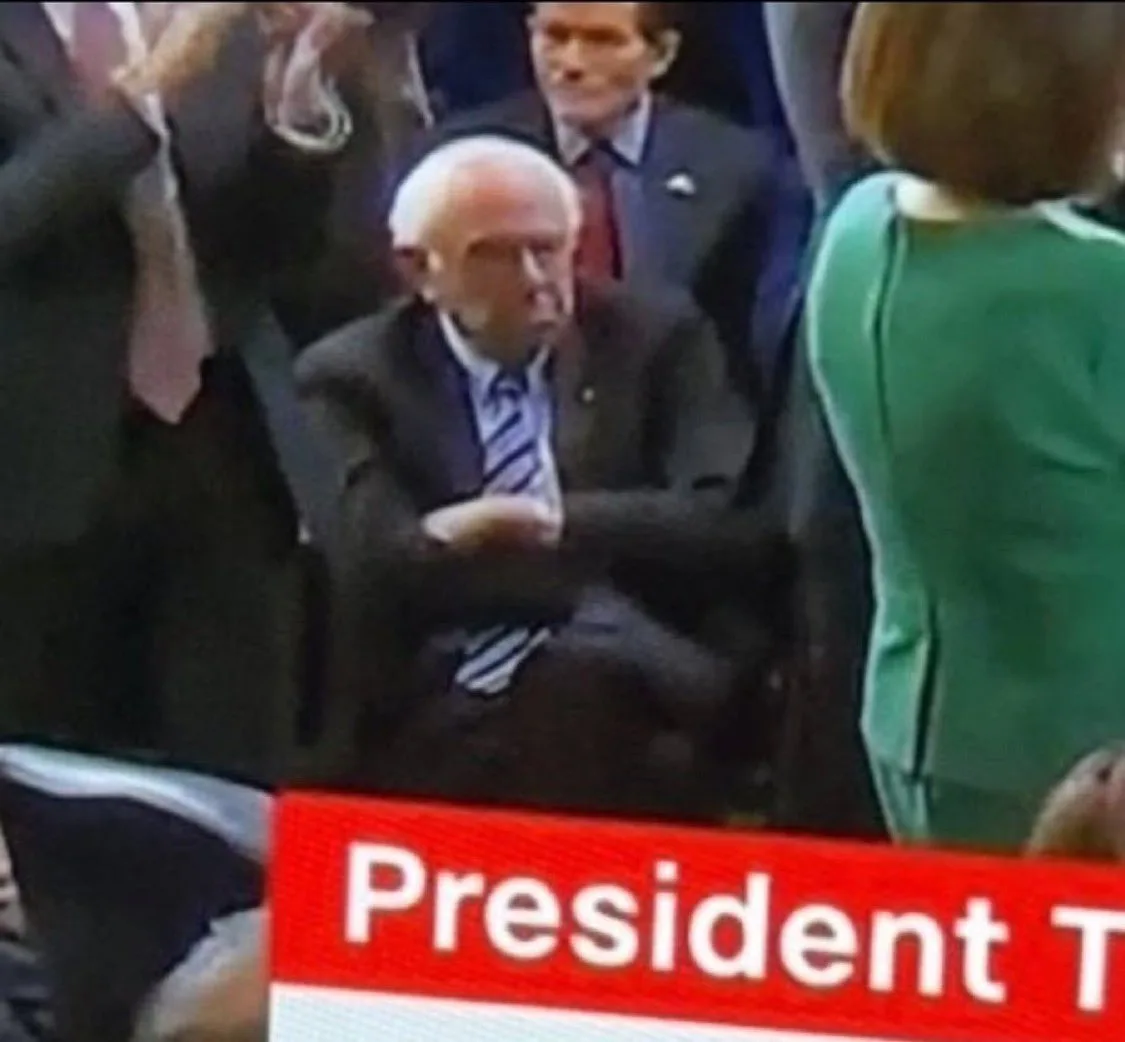 A low-resolution image shows an older man in a dark suit and striped tie seated, looking slightly to his right with a…