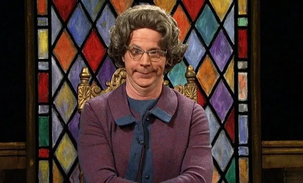 A man with curly gray hair, glasses, and a purple cardigan sits in a golden throne in front of a colorful stained-glass…