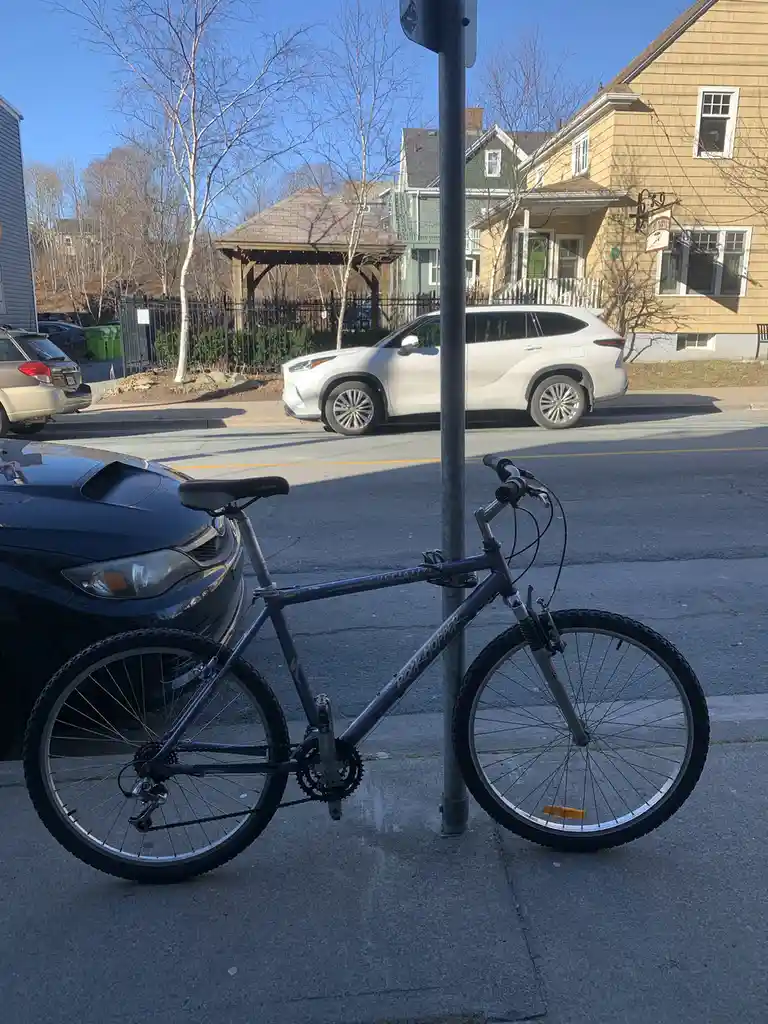 A dark blue mountain bike is locked to a metal pole on a sidewalk next to a parked black car. Across the street, a white SUV…