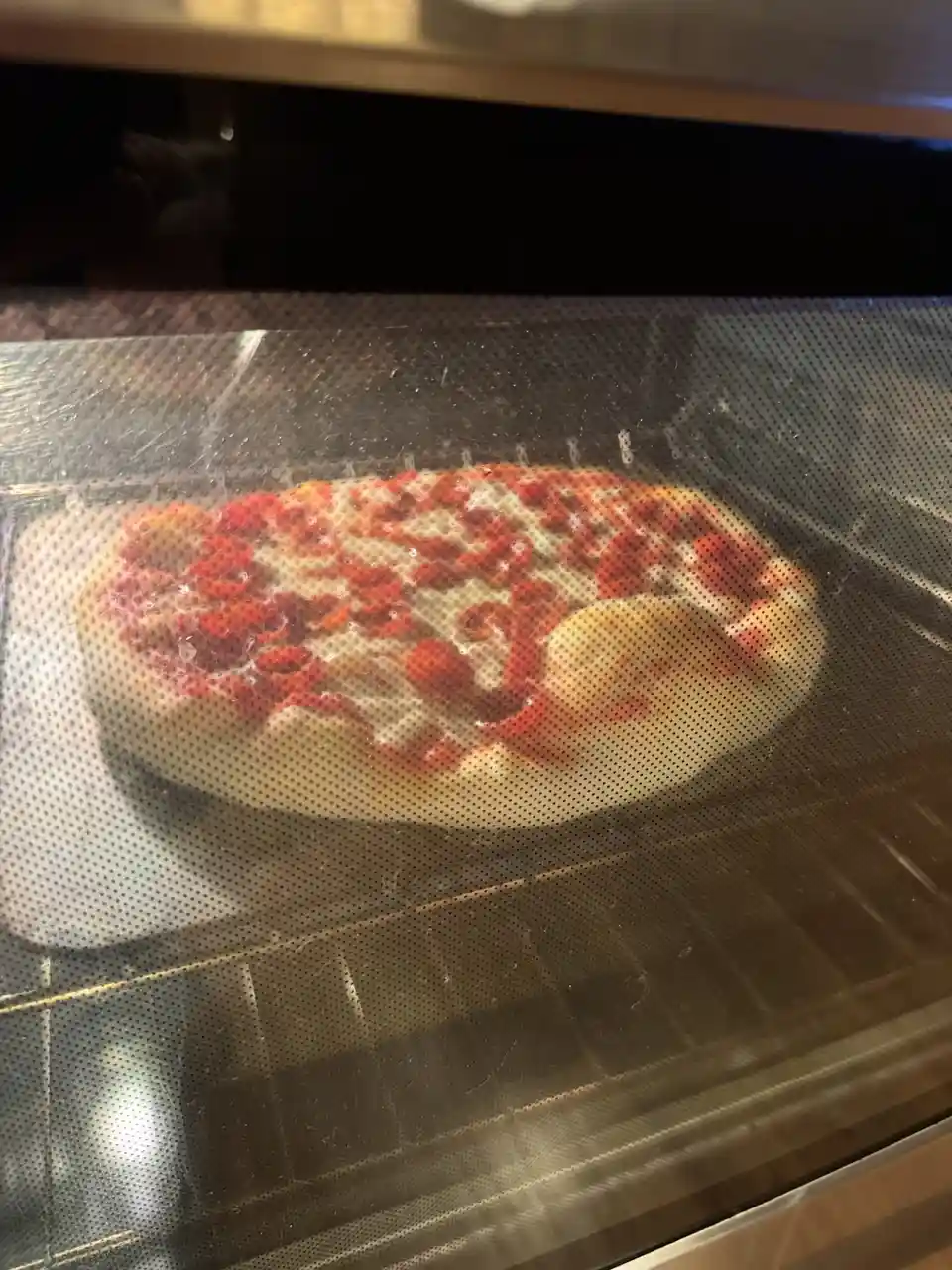 A pizza inside an oven, viewed through a perforated glass door, has a large, perfectly round, uncooked-looking object on one…