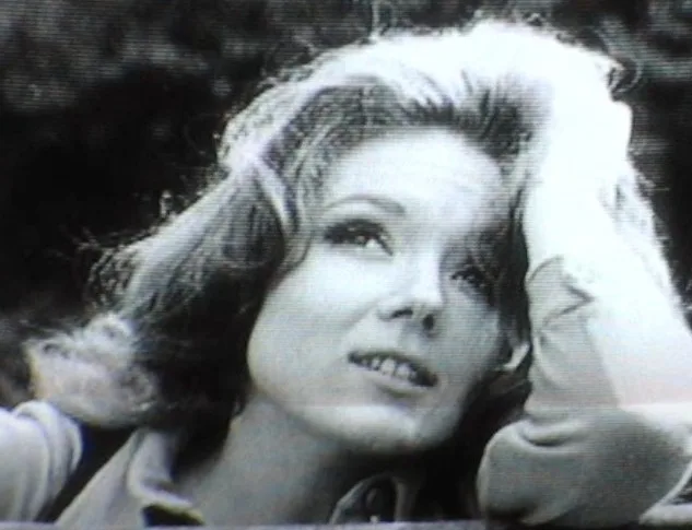 A grainy black-and-white photo shows a woman with wavy hair looking upward with a slightly amused or resigned expression…