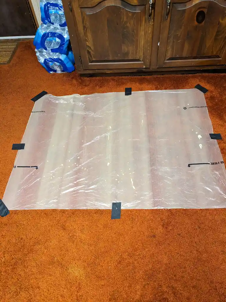 A large, clear plastic sheet, likely from a moving or storage box, is taped down on an orange carpet with black tape at its…