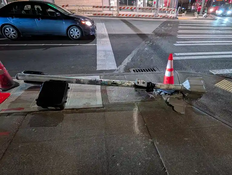 A broken, knocked-over crosswalk signal pole lies on a sidewalk at night, with a red and white traffic cone nearby and a…