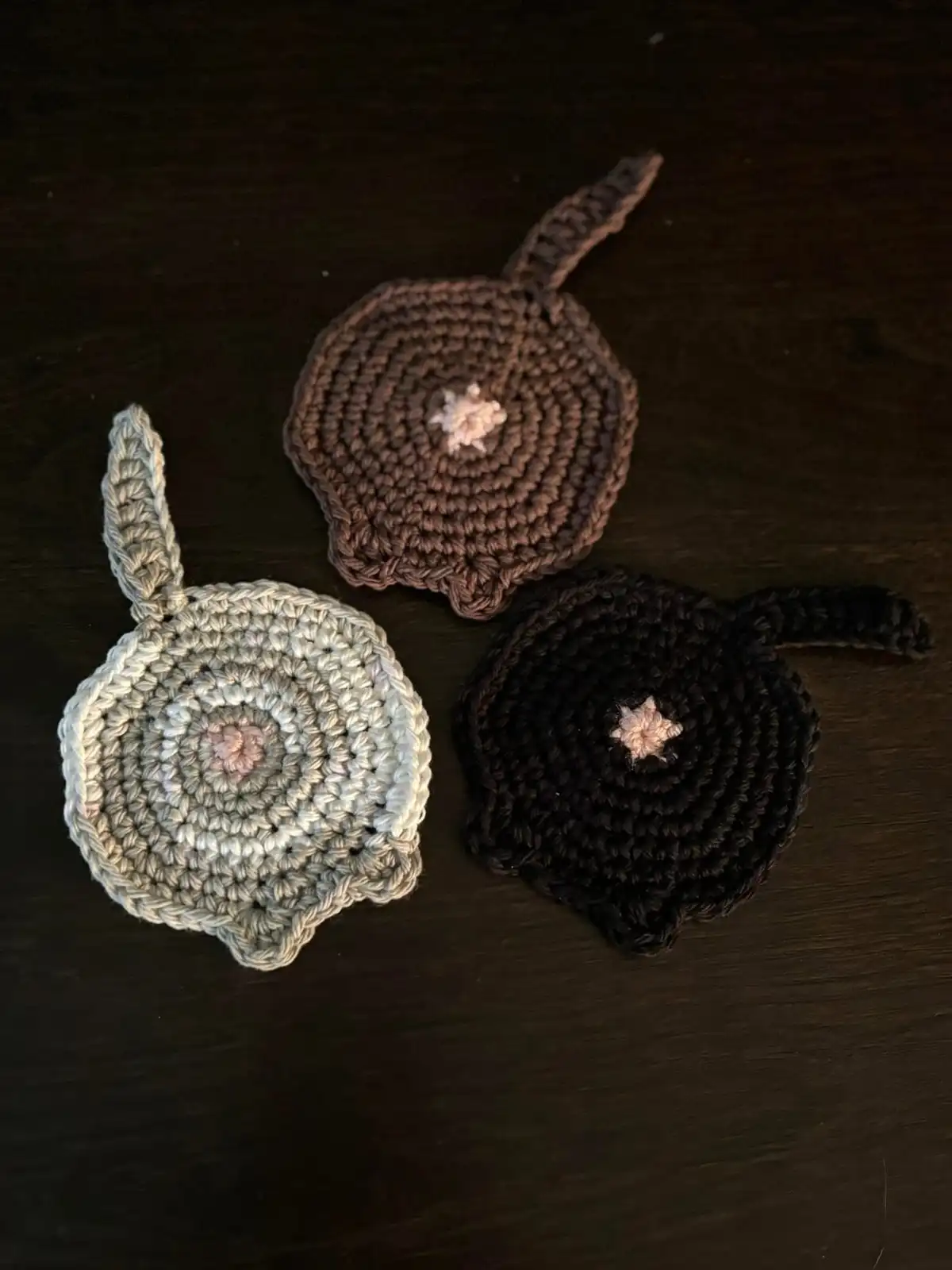 Three hand-crocheted, circular, apple-shaped items in brown, gray, and black with pink centers and yarn tails lie on a dark…