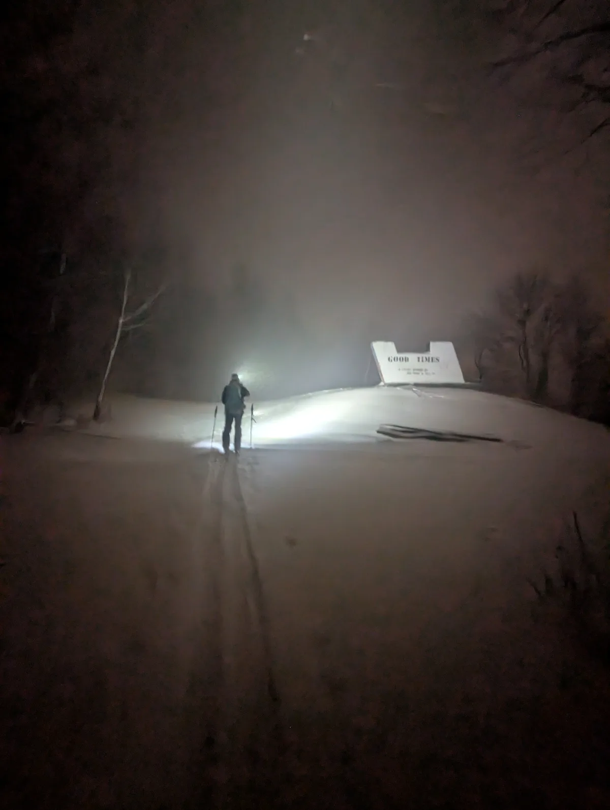 A person is cross-country skiing at night in deep snow, illuminated by a headlamp, approaching a large white sign that reads…