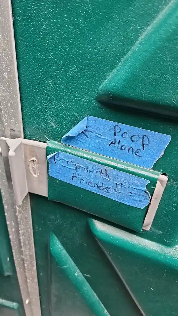 Two pieces of blue painter's tape are stuck to a green plastic door hinge. One tape reads 'Poop Alone' with an arrow…