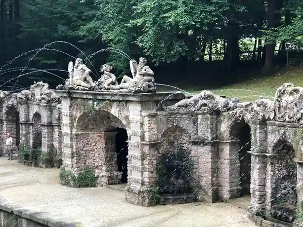 An ornate, weathered stone fountain with multiple arches and statues of mythological figures, water spraying from various…