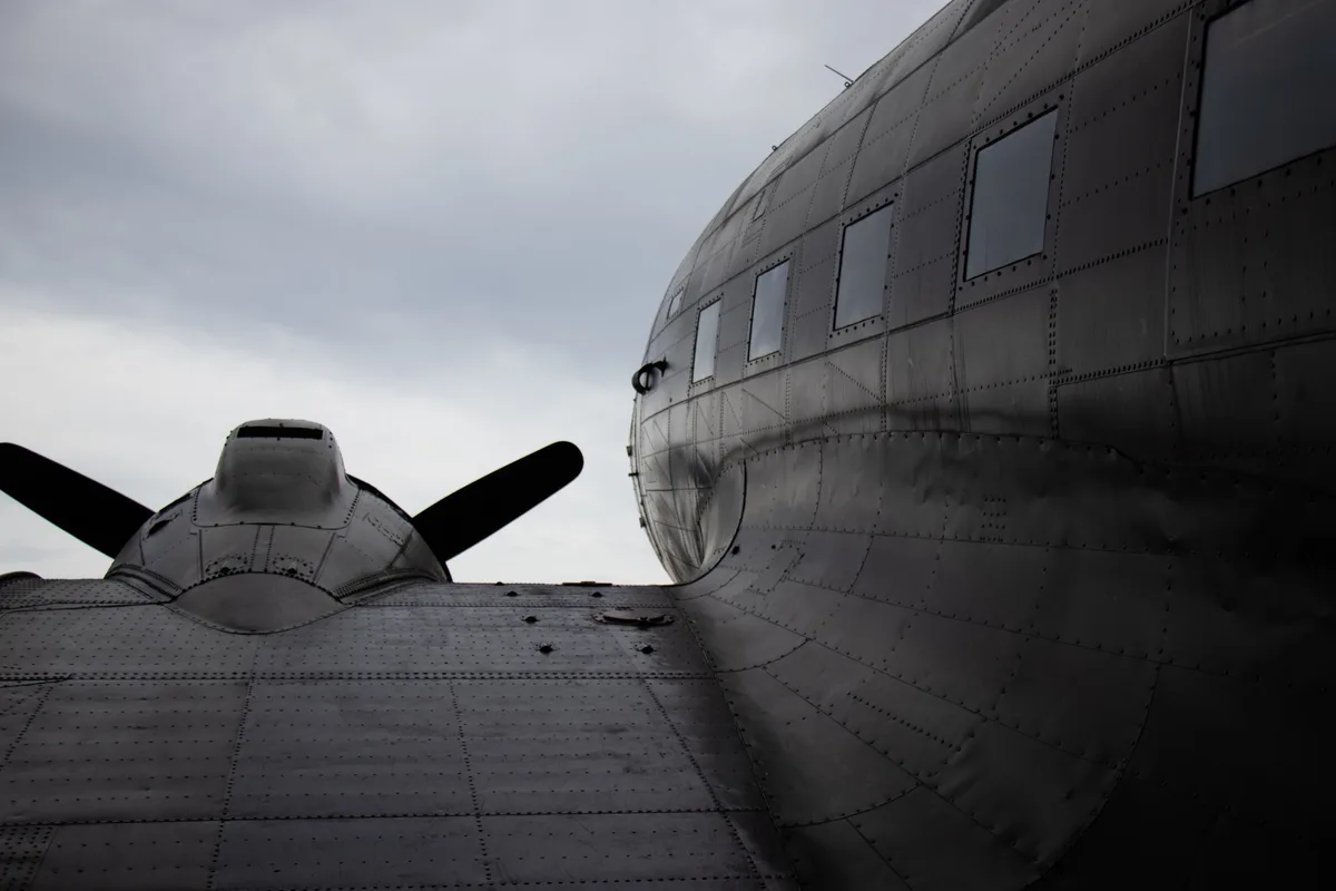 A low-angle black-and-white photo shows the nose and cockpit of a vintage propeller airplane on the left, facing the curved…