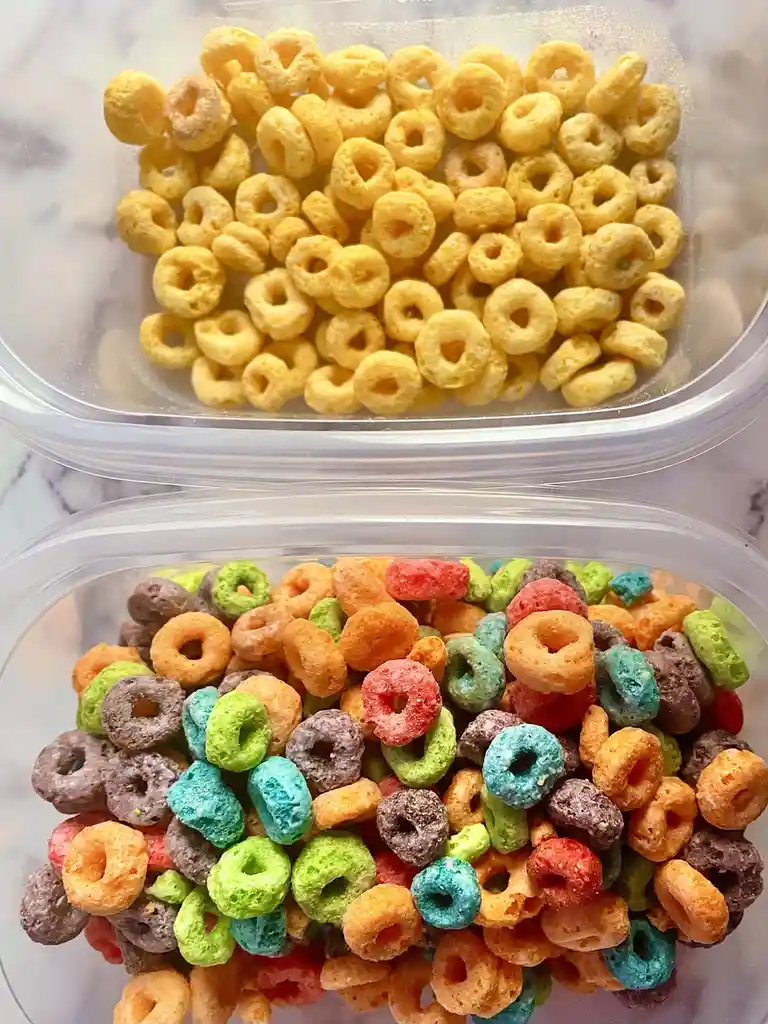 Two clear plastic containers sit side by side, one filled with plain yellow cereal loops and the other with colorful…