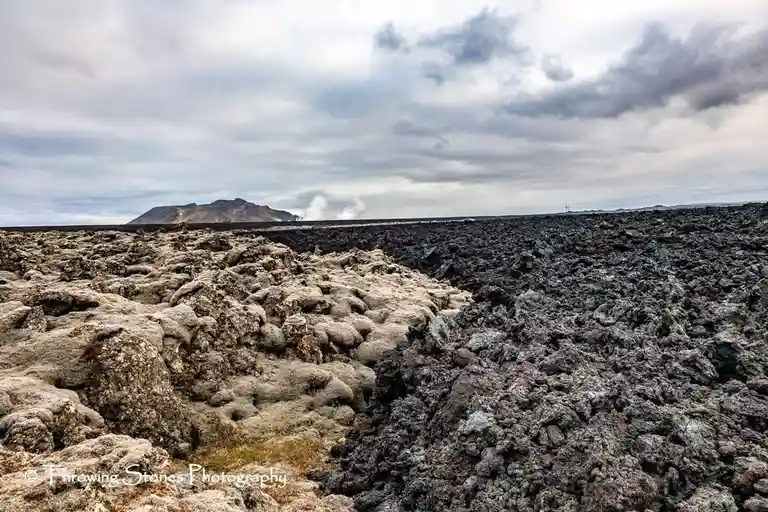 The image shows a wide, desolate lava field with dark, jagged rock formations in the foreground and a distant mountain…