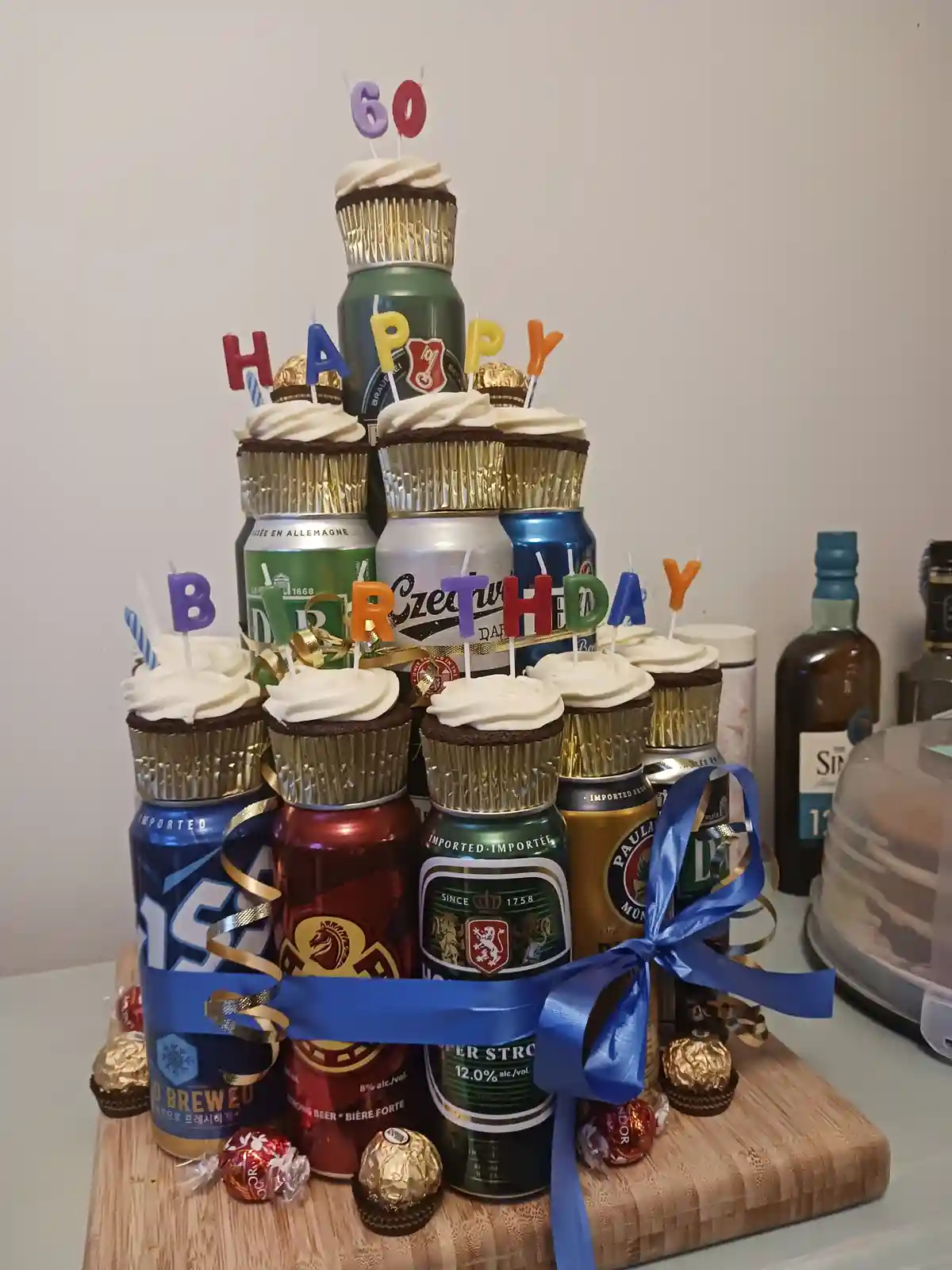 A tiered birthday cake made of stacked beer cans with chocolate cupcakes placed on top of each can, decorated with colorful…