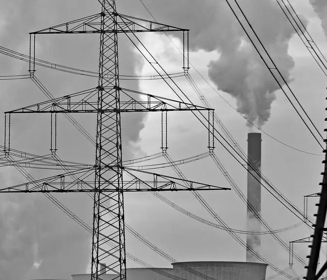 A black-and-white photo shows a tall electricity pylon with many wires against a cloudy sky, with a smokestack emitting…