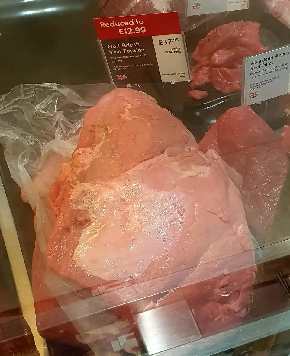 A large, pale pink, uncooked meat cut sits in a supermarket meat display case, with a red sign above it reading 'Reduced to…