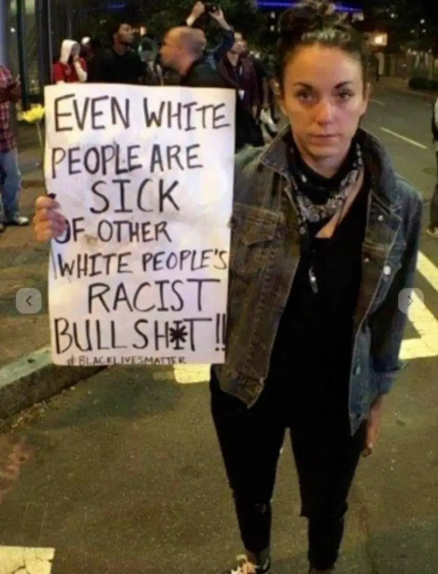 A woman stands outdoors at night holding a handmade protest sign that reads 'EVEN WHITE PEOPLE ARE SICK OF OTHER WHITE…