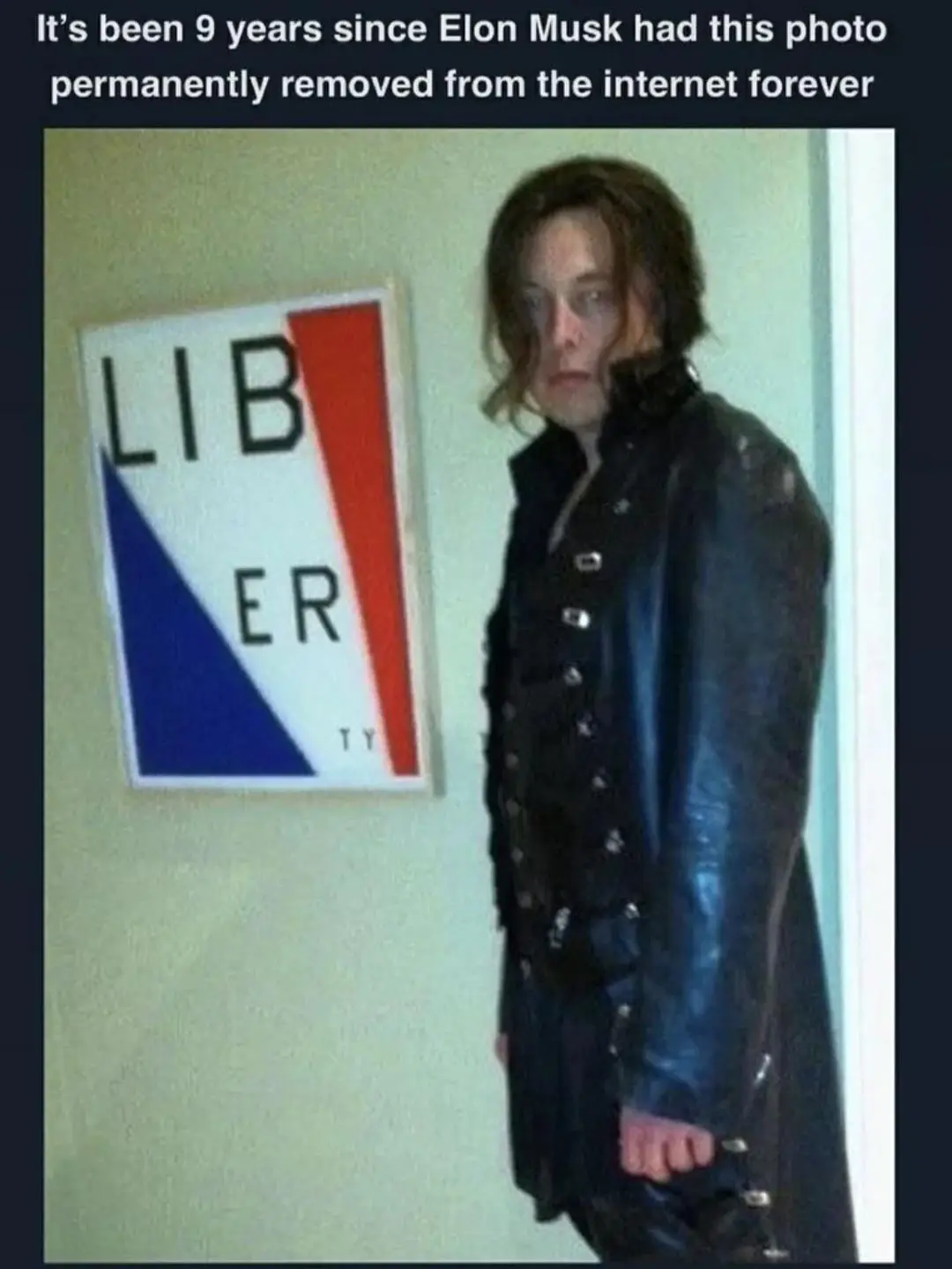 A person with shoulder-length brown hair and a black leather coat stands beside a framed sign that reads 'LIBERTY' in black…