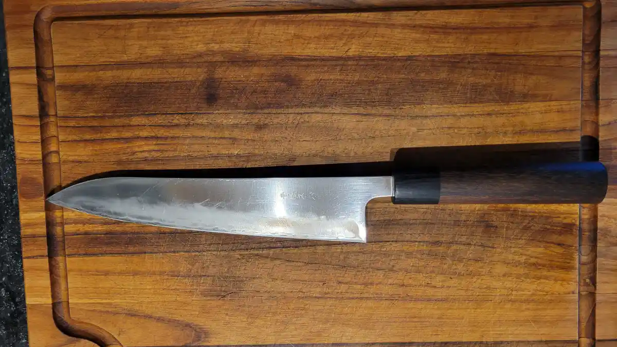 A chef's knife with a dark wooden handle and metallic blade rests horizontally on a well-used wooden cutting board. The…