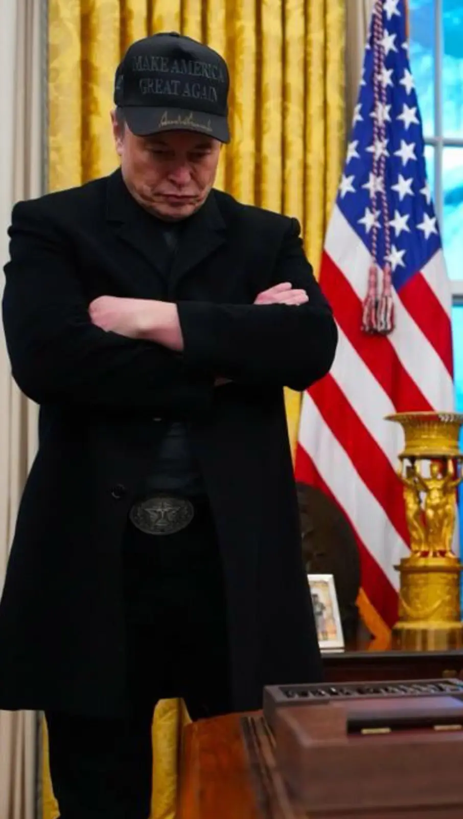 A man in a black coat and a 'Make America Great Again' cap stands with arms crossed in front of an American flag and golden…