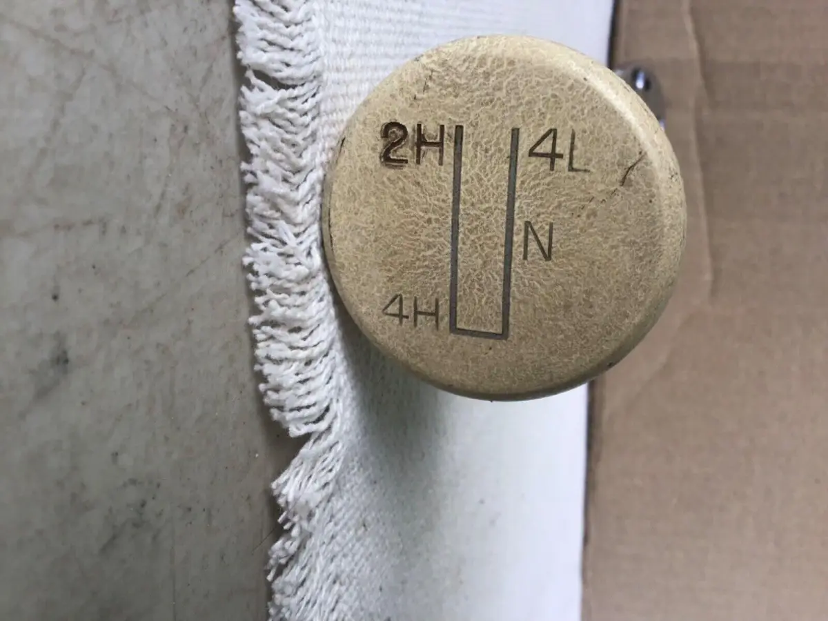 A close-up of a beige, textured gear selector knob with engraved positions: 2H, 4L, N, and 4H, arranged in a U-shape. The…